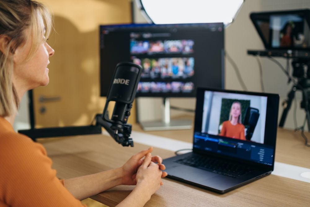 Eine Mitarbeiterin sitzt vor einem Laptop, Bildschirm und Mirkofon und nimmt etwas auf.