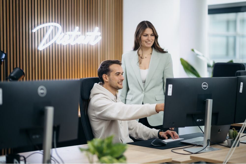 Ein männlicher Mitarbeiter sitzt an seinem Schreibtisch in einem modernen Büro und zeigt einer weiblichen Mitarbeiterin etwas auf seinem Bildschirm. Das Distart-Logo leuchtet im Hintergrund.