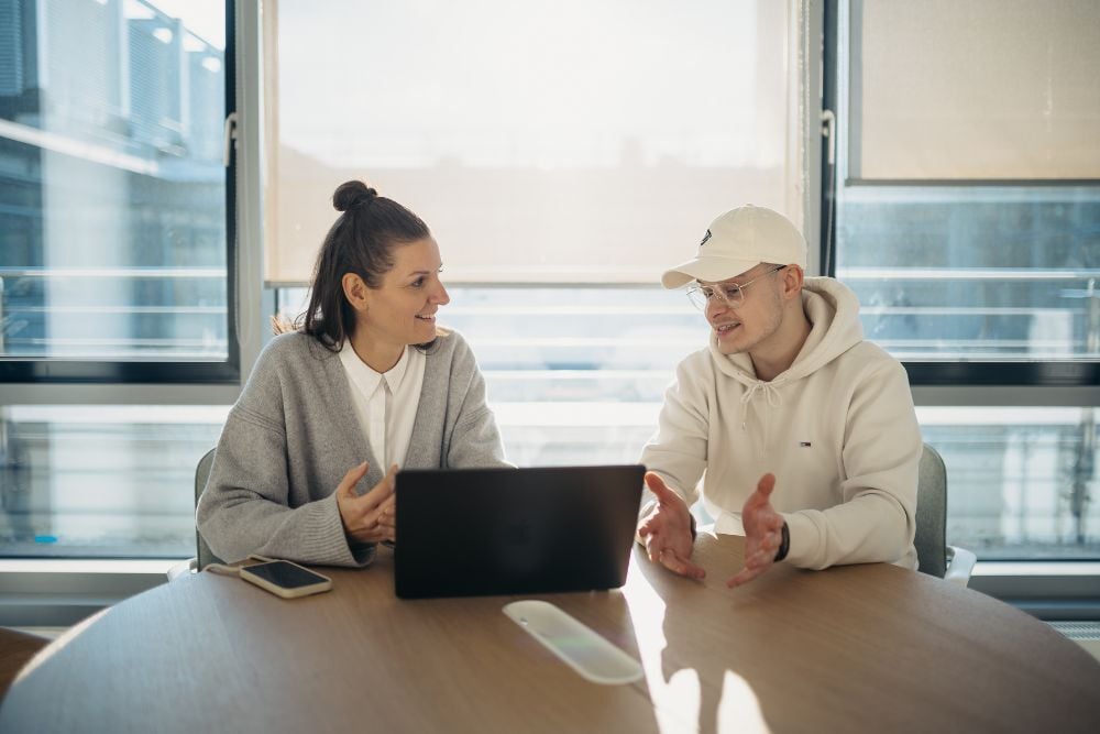 Zwei Mitarbeitende besprechen sich gut gelaunt an einem Tisch, vor Ihnen ein aufgeklappter Laptop. die sonne scheint in das Zimmer.