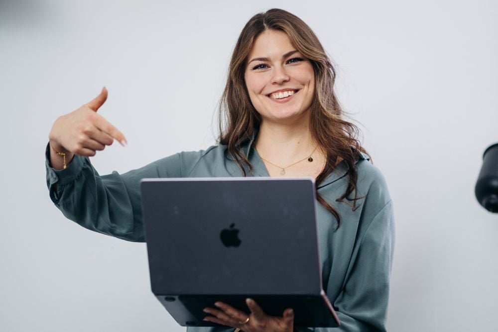 Eine Distart Mitarbeoiterin hält ihren silbernen MacBook mit einer Hand und zeigt lächelnd mit der anderen Hand darauf.