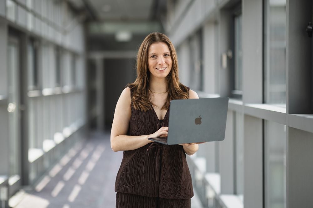 Eine Distart Mitarbeiterin steht in einer hellen Umgebung, lächelt und hält einen Laptop in den Händen