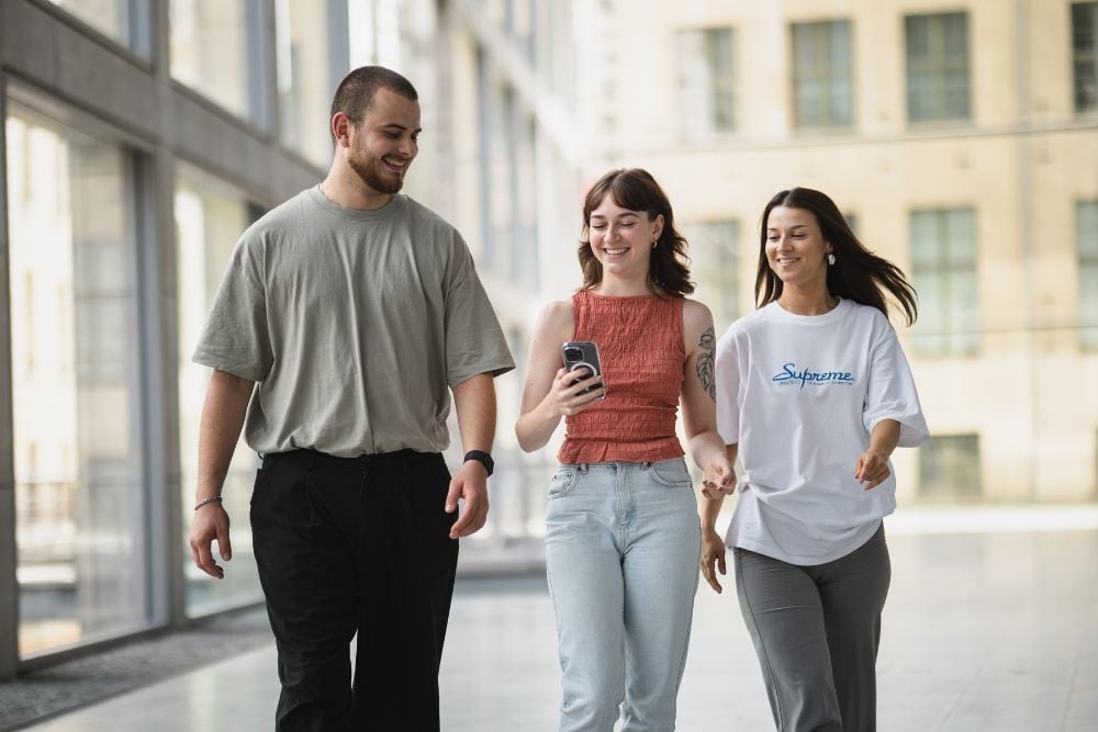 Drei junge Menschen gehen durch ein lichtdurchflutetes Gebäude. Alle lächeln und scheinen begeistert über etwas, dass die mittlere Person auf ihrem Smartphone anzeigt.